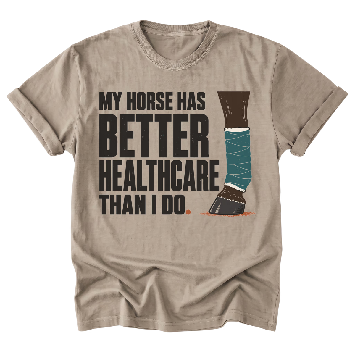 A beige sand crewneck t-shirt with a graphic print that reads 'MY HORSE HAS BETTER HEALTHCARE THAN I DO' alongside an illustration of an equestrian leg wrapped in teal bandages. The t-shirt is displayed flat against a solid white background.