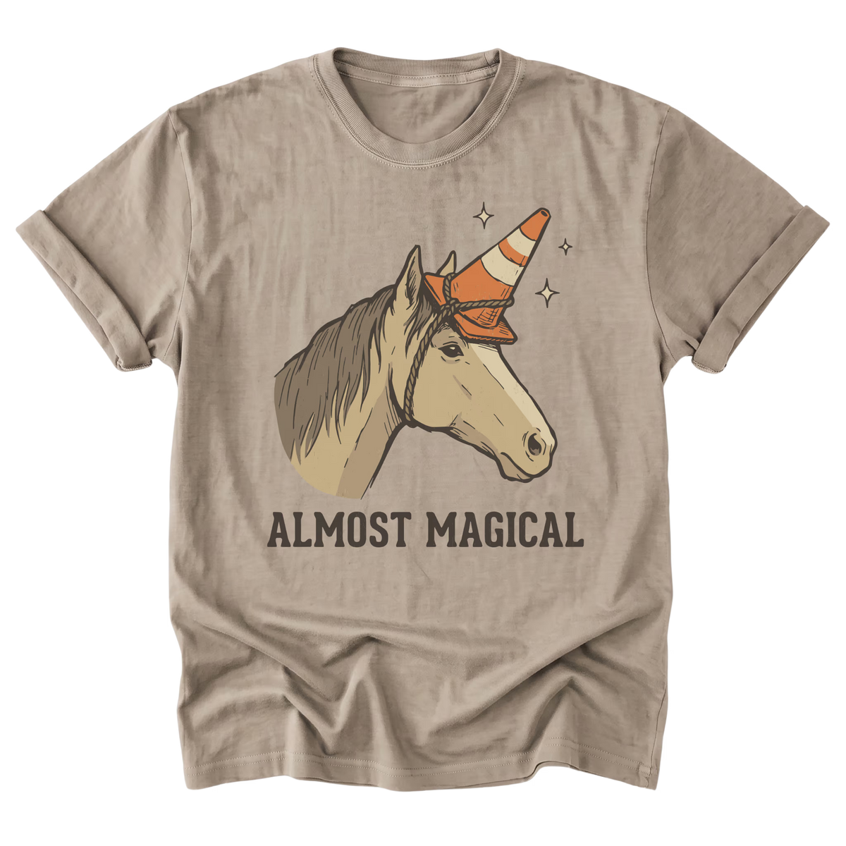 A sand beige graphic t-shirt featuring an illustration of a horse with a traffic cone horn and the text 'ALMOST MAGICAL' printed below it, displayed on a clean white background.